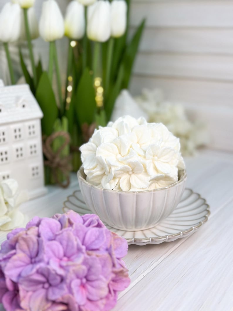 May include: A still-life composition featuring a white ceramic bowl filled with white flowers, resting on a matching saucer. A purple hydrangea and white tulips are in the background, with a small white house. The scene is set on a white wooden surface.