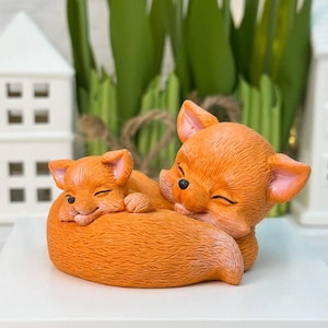 Peut inclure: Figurine en céramique représentant deux renards orange, l'un niché contre l'autre. Les renards ont les yeux fermés et sont enroulés l'un contre l'autre. La figurine est sur une surface blanche, avec un fond de verdure verte et de maisons décoratives blanches.