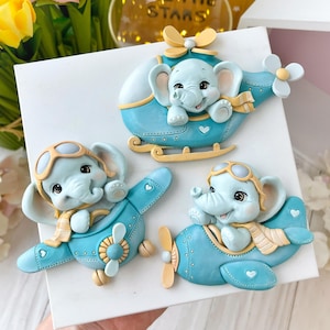 May include: Three blue and yellow airplane and helicopter figurines with elephant pilots. Each elephant is wearing a scarf and goggles. The figurines are on a white surface.