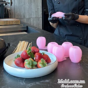 Pu&ograve; includere: Fragole fresche con gambi verdi su un piatto, accanto a stampi rosa e spiedini di legno. Una persona in uniforme nera e guanti neri tiene in mano uno stampo rosa. Il testo "Mold.com.ua Orehova Cakes" &egrave; visibile.