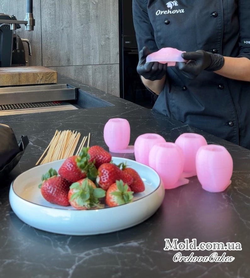 Pu&ograve; includere: Fragole fresche con gambi verdi su un piatto, accanto a stampi rosa e spiedini di legno. Una persona in uniforme nera e guanti neri tiene in mano uno stampo rosa. Il testo "Mold.com.ua Orehova Cakes" &egrave; visibile.
