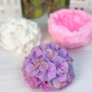 May include: A close-up of three floral-shaped soaps. The front soap is a lavender hydrangea with gold accents. Behind it is a white soap and a pink peony soap. All soaps are on a white wooden surface.