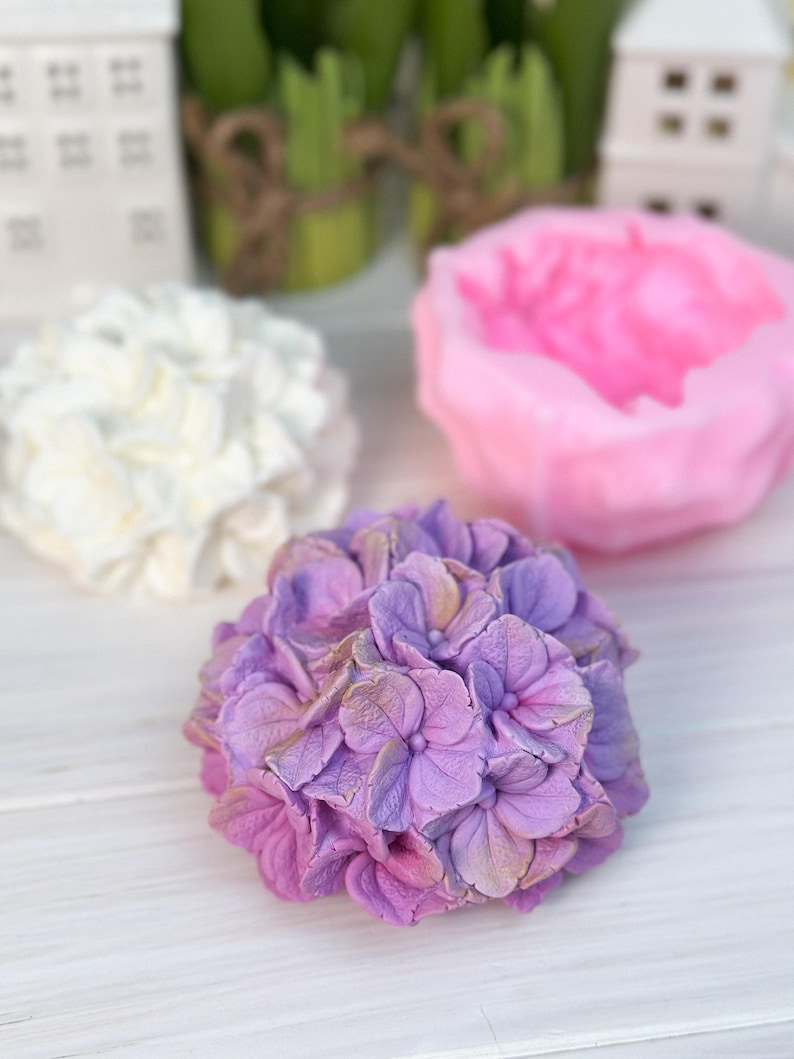 May include: A close-up of three floral-shaped soaps. The front soap is a lavender hydrangea with gold accents. Behind it is a white soap and a pink peony soap. All soaps are on a white wooden surface.