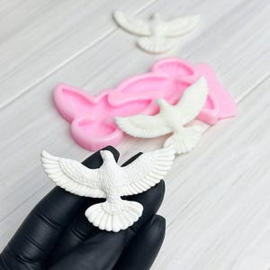 Peut inclure: Décorations blanches en forme de colombe aux ailes déployées. Un moule en silicone rose est visible en arrière-plan. Les figurines de colombes sont probablement destinées à l'artisanat ou à la décoration de gâteaux.