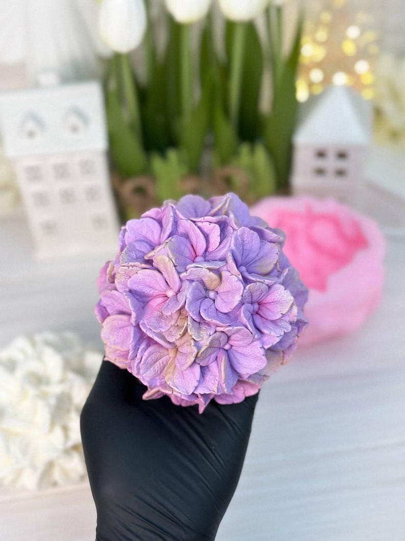 May include: A handcrafted, lavender-colored hydrangea flower with a detailed petal design. The flower is held in a black glove, with a blurred background featuring white tulips and decorative houses. The flower's petals have a gradient of pink and gold.