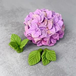 May include: A sculpted hydrangea flower head in shades of light purple and pink, accompanied by three separate green leaves with visible vein textures. The flower petals are layered and rounded, creating a dense, spherical cluster of blossoms.