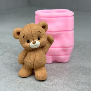 Op de afbeelding: Een bruin teddybeerbeeldje met een glimlachende uitdrukking en een opgeheven arm. De beer heeft zwarte ogen en een witte snuit. Op de achtergrond staat een roze siliconen mal.