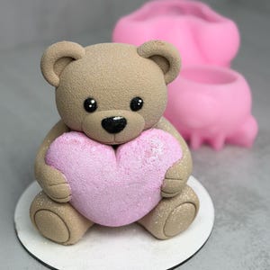 May include: A tan teddy bear figurine holding a pink heart. The bear has black eyes and a black nose. The heart is covered in glitter. A pink mold is in the background. The bear is sitting on a white circular base.