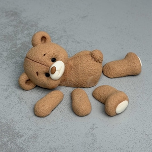 Puede incluir: Figura de oso de peluche marrón desmontada, con cara y patas blancas. La cabeza, el cuerpo y las extremidades separadas están sobre una superficie gris. El oso tiene ojos negros y nariz marrón.