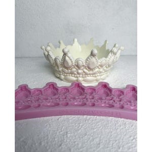 May include: A white crown mould with a pink silicone mould of a crown with a beaded design. The silicone mould has the text "КОРОНА ДИАМ. 100мм" on it.