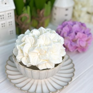 May include: A white hydrangea flower arrangement in a decorative, scalloped-edge bowl and matching plate. The bowl and plate are a light beige color with a silver trim. A purple hydrangea and small white houses are in the background.