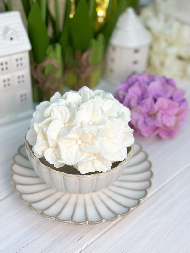 May include: A white hydrangea flower arrangement in a decorative, scalloped-edge bowl and matching plate. The bowl and plate are a light beige color with a silver trim. A purple hydrangea and small white houses are in the background.