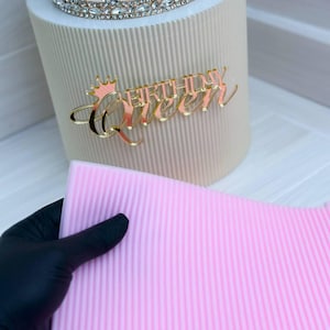 May include: Pink cake decorating mat with raised lines. The mat is made of silicone and is designed to help create smooth, even frosting on cakes. The mat is shown on top of a white cake with a gold crown and the words "Birthday Queen" on top.