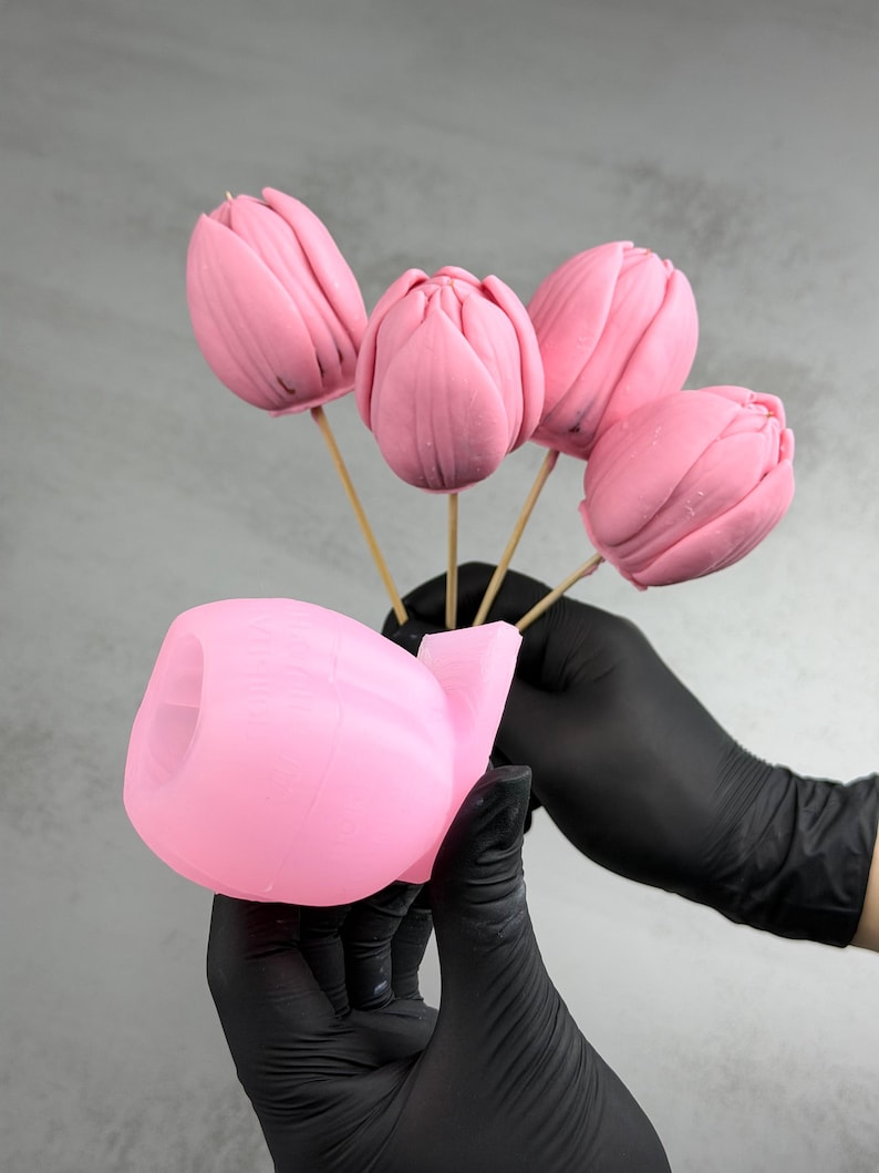 Pu&ograve; includere: Cinque dolcetti rosa a forma di tulipano su bastoncini di legno, tenuti sopra uno stampo rosa. I dolcetti hanno una finitura liscia e lucida. Lo sfondo &egrave; grigio chiaro. I dolcetti sono tenuti da una persona con guanti neri.