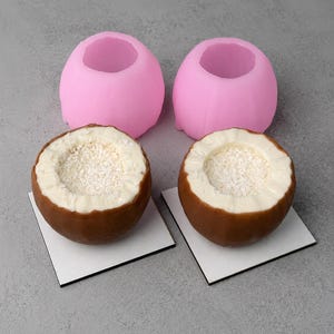 May include: Two pink silicone molds positioned behind two brown chocolate shells filled with white cream and shredded coconut. Each chocolate shell rests on a small square white base, showcasing a hollow center filled with white coconut flakes.