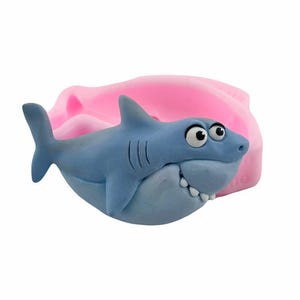 May include: A blue cartoon shark with a wide grin and white teeth. The shark has large, expressive eyes and is set against a pink background. The shark is a fun, whimsical design.