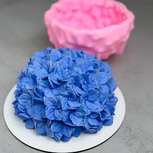 Op de afbeelding: Een blauwe hortensia-vormige cake op een wit bord, met een roze mal op de achtergrond. De cake is bedekt met gedetailleerde blauwe bloemblaadjes, waardoor een textuurrijk, bloemig ontwerp ontstaat. De roze mal heeft een vergelijkbaar bloemmotief.