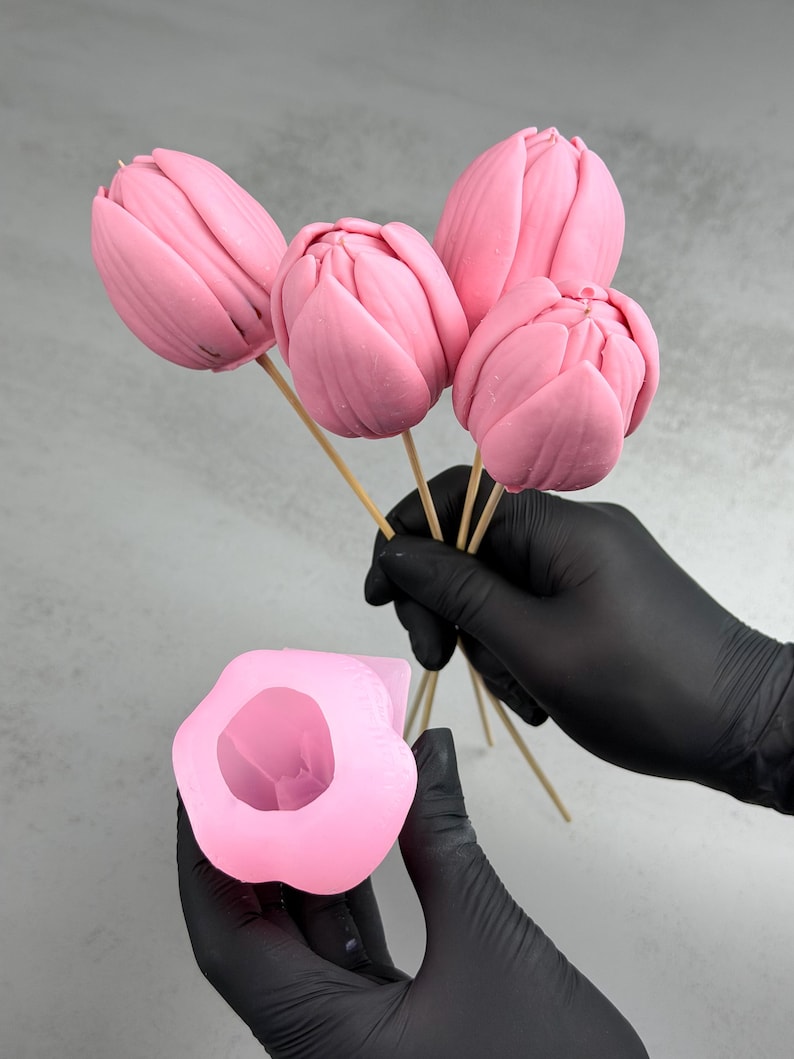 Pu&ograve; includere: Fiori di tulipano rosa su spiedini di legno, tenuti da mani guantate, accanto a uno stampo rosa a forma di fiore. I tulipani hanno una consistenza liscia e sono di una tonalit&agrave; rosa vibrante. Lo stampo &egrave; anch'esso rosa e ha la forma di un fiore.