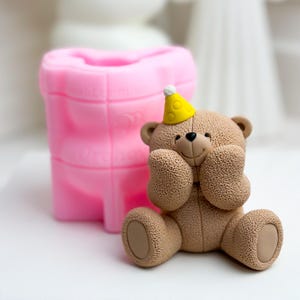 May include: A pink silicone mold shaped like a teddy bear wearing a yellow party hat. The mold is used to create a cake topper or other edible decorations.