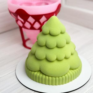 May include: A light green, tiered, cone-shaped dessert with a smooth, matte texture, resting on a circular white base. Behind it sits a pink and red silicone mold with a diamond-patterned cutout design, used for shaping such confections.