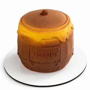 May include: A brown, barrel-shaped dessert with a lid, resembling a honey pot. The word "HONEY" is displayed on the front. The top of the dessert has a yellow, honey-like substance dripping down the sides. The dessert sits on a white, round plate.