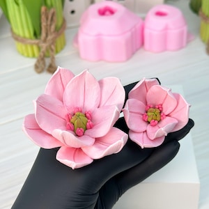 May include: Two pink, handcrafted magnolia flowers with green and pink centers. The flowers are held in a black gloved hand, with a white block in the background. Other pink flower molds and green plants are also visible.
