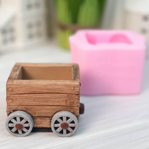 Puede incluir: Una maceta con forma de carro de textura madera marrón con cuatro ruedas grises con centros marrones, situada frente a un molde de silicona rosa suave. El carro tiene una parte superior rectangular abierta y detalles de tablones horizontales.
