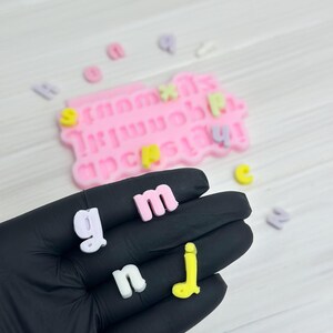 Puede incluir: Una mano sosteniendo una variedad de letras de colores, incluyendo rosa, blanco, amarillo y morado. Las letras est&aacute;n hechas de un material suave y flexible y probablemente se usan para manualidades o decoraci&oacute;n.