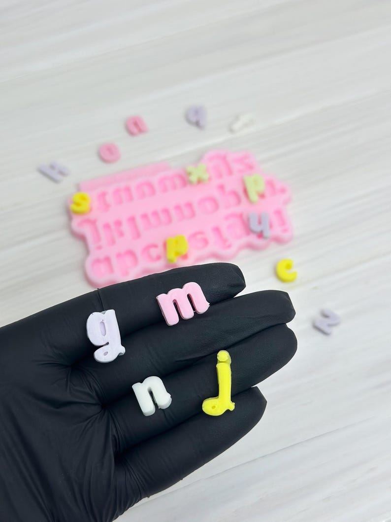 Puede incluir: Una mano sosteniendo una variedad de letras de colores, incluyendo rosa, blanco, amarillo y morado. Las letras est&aacute;n hechas de un material suave y flexible y probablemente se usan para manualidades o decoraci&oacute;n.