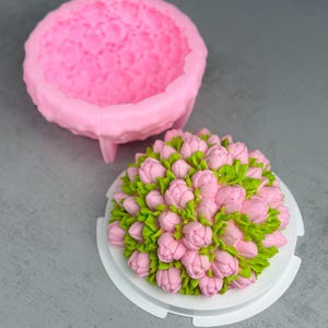 May include: A pink silicone mold and a cake decorated with pink buttercream flowers and green leaves. The cake is on a white round base, and the mold is a round shape with a textured interior.