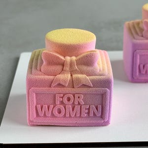 Può includere: Una torta a forma di flacone di profumo rosa e giallo sfumato, con un fiocco e le parole "FOR WOMEN" in rilievo sul davanti. La torta ha un aspetto strutturato e vellutato e poggia su una superficie bianca.
