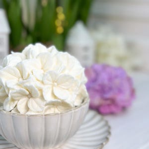 May include: A white ceramic bowl filled with white flower-shaped decorations, sitting on a matching saucer. The bowl and saucer have a ribbed design. A purple flower and other decorations are in the background.