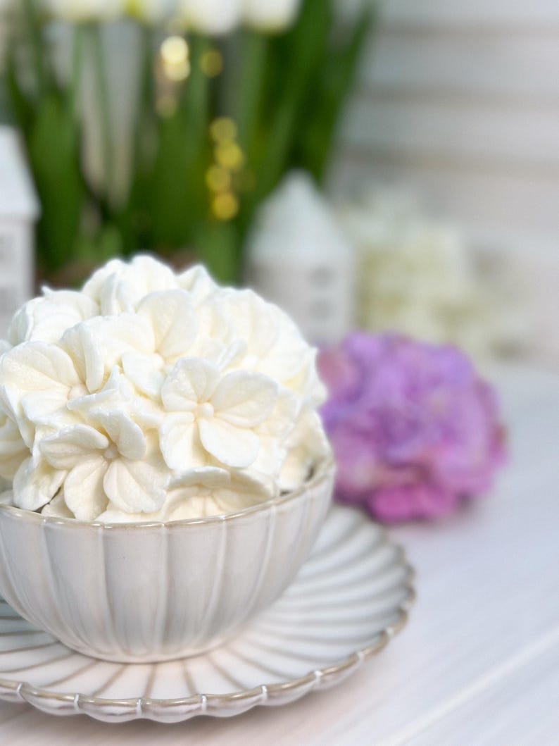May include: A white ceramic bowl filled with white flower-shaped decorations, sitting on a matching saucer. The bowl and saucer have a ribbed design. A purple flower and other decorations are in the background.