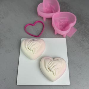 May include: A selection of heart-shaped moulds and a cookie cutter. Two pink moulds with legs and a heart-shaped cookie cutter are visible. Two heart-shaped soaps with clasped hands are on a white surface.