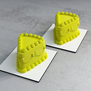 Puede incluir: Dos porciones de pastel verde lima con glaseado decorativo, cada una sobre una base cuadrada blanca con borde negro. Los pasteles tienen una superficie texturizada y están decorados con un borde festoneado y detalles ornamentados.