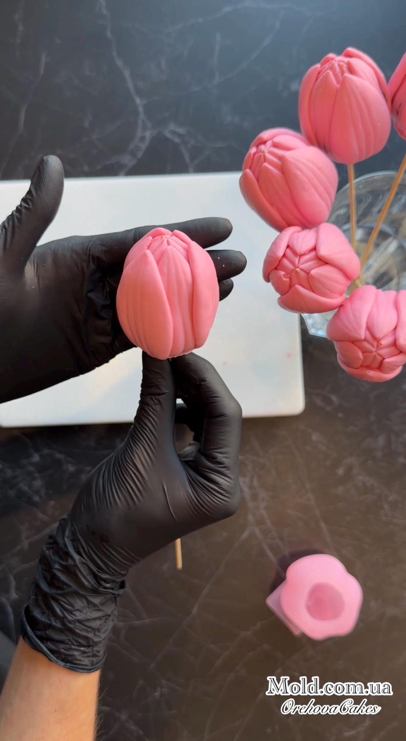 Pu&ograve; includere: Dolci rosa a forma di tulipano su bastoncini di legno, forse cake pops o caramelle. Uno &egrave; tenuto da una mano guantata, altri sono disposti in un bicchiere. &Egrave; visibile anche uno stampo a forma di fiore.