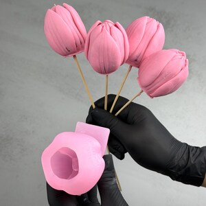 Pu&ograve; includere: Cinque caramelle rosa a forma di tulipano su bastoncini di legno. Le caramelle sono tenute da una persona con guanti neri, insieme a uno stampo in silicone rosa. Le caramelle hanno una consistenza liscia e sono di un rosa vibrante.