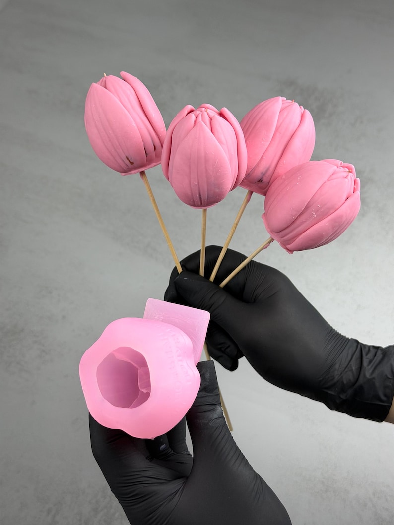 Pu&ograve; includere: Cinque caramelle rosa a forma di tulipano su bastoncini di legno. Le caramelle sono tenute da una persona con guanti neri, insieme a uno stampo in silicone rosa. Le caramelle hanno una consistenza liscia e sono di un rosa vibrante.