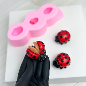 May include: Three red and black ladybug figurines with black dots. One ladybug is open with a brown interior. The ladybugs are on a white surface next to a pink silicone mold with three ladybug-shaped cavities.