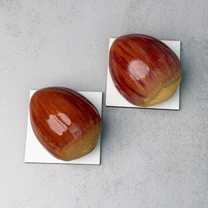 May include: Two glossy, brown, acorn-shaped objects with textured, tan-colored bases. Each acorn is mounted on a small, square, white base. The surfaces of the acorns feature vertical, darker brown streaks and a smooth, reflective finish.