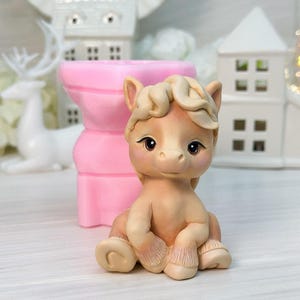 Puede incluir: Una figura de poni de color canela claro con una melena rizada y ojos oscuros se sienta sobre una superficie blanca. Un molde rosa está detrás del poni. Casas decorativas blancas y un ciervo en el fondo.