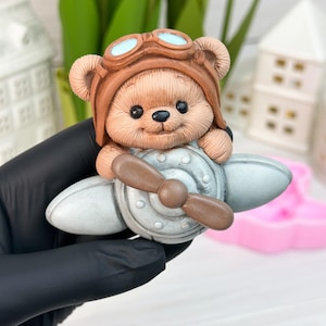 May include: A brown teddy bear figurine wearing a brown aviator helmet and goggles, sitting in a light grey airplane with a brown propeller. The bear has a smiling expression. A whimsical decorative item.