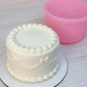Puede incluir: Un pastel blanco glaseado con glaseado decorativo en los bordes superior e inferior, y un diseño de guirnalda. El pastel está sobre una base blanca. Un cuenco rosa está en el fondo.