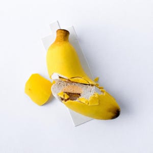 May include: A banana-shaped dessert with a bright yellow outer shell that is cracked open to reveal a light-colored interior filling and a darker center. The fruit-shaped treat features brown tips and rests on a small, rectangular white base.