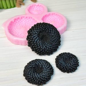 Può includere: L'immagine mostra uno stampo in silicone rosa con tre cavità a forma di fiore, insieme a tre saponi neri strutturati a forma di fiore. I saponi hanno un design dettagliato e stratificato, disposti su una superficie di legno bianco.
