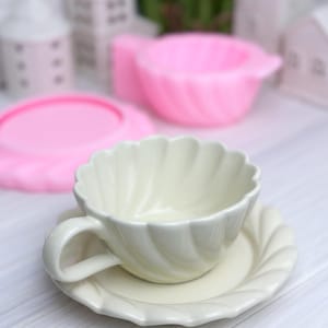 Könnte beinhalten: Ein cremefarbenes Tee-Set mit gewelltem Rand. Die Tasse hat einen gebogenen Henkel und steht auf einer passenden Untertasse. Im Hintergrund befinden sich rosa Formen und Miniatur-Weißhäuser.