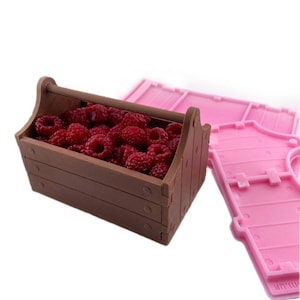 May include: A brown wooden toolbox filled with red raspberries. The toolbox has a handle on top and is made of a moldable material. The mold is pink and is shown next to the toolbox.