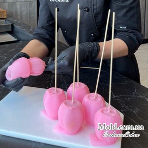 Pu&ograve; includere: Mele caramellate rosa su bastoncini, presentate su un tagliere bianco. Le mele hanno una finitura liscia e lucida e sono disposte in fila. L'immagine mostra anche una persona con guanti neri che tiene in mano altre mele caramellate rosa.