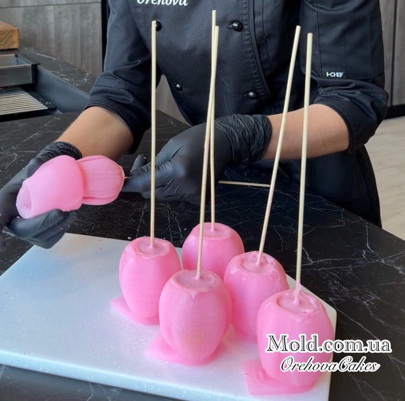 Pu&ograve; includere: Mele caramellate rosa su bastoncini, presentate su un tagliere bianco. Le mele hanno una finitura liscia e lucida e sono disposte in fila. L'immagine mostra anche una persona con guanti neri che tiene in mano altre mele caramellate rosa.