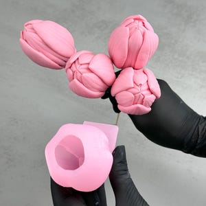 Pu&ograve; includere: Stampo in silicone a forma di tulipano rosa e un bouquet di boccioli di tulipano rosa. Lo stampo &egrave; tenuto da una persona con guanti neri. I boccioli di tulipano sono su uno stelo di legno. Lo sfondo &egrave; di colore grigio chiaro.